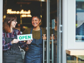 Two woman holding an open sign in front of a store.