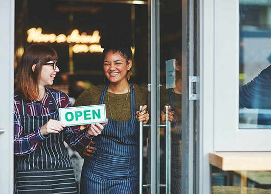 Two woman holding an open sign in front of a store.