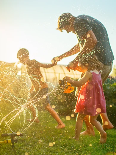 A father and two kids playing with the backyard sprinklers