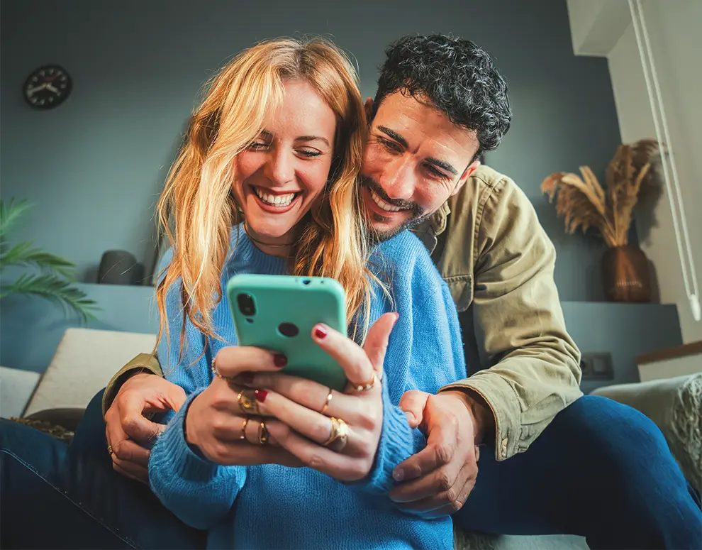 Couple on couch smiling at smartphone, cozy home setting.