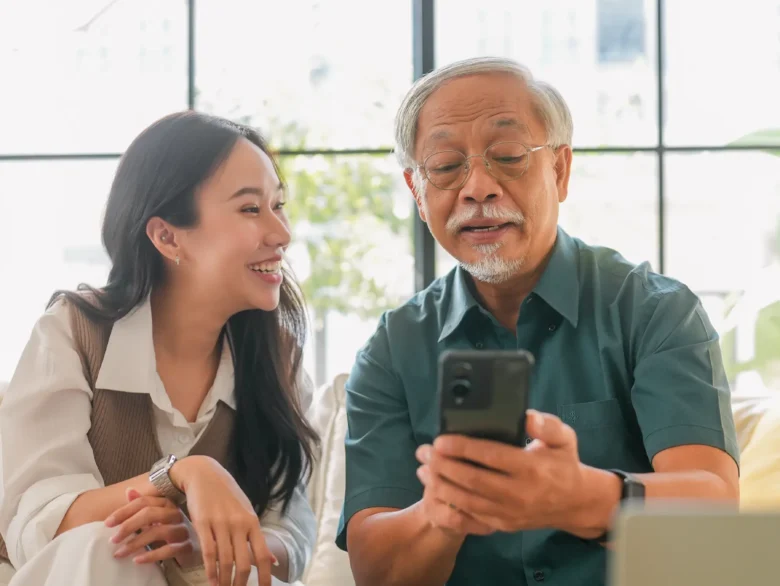 An adult woman smiling at an elderly man as he uses a phone.