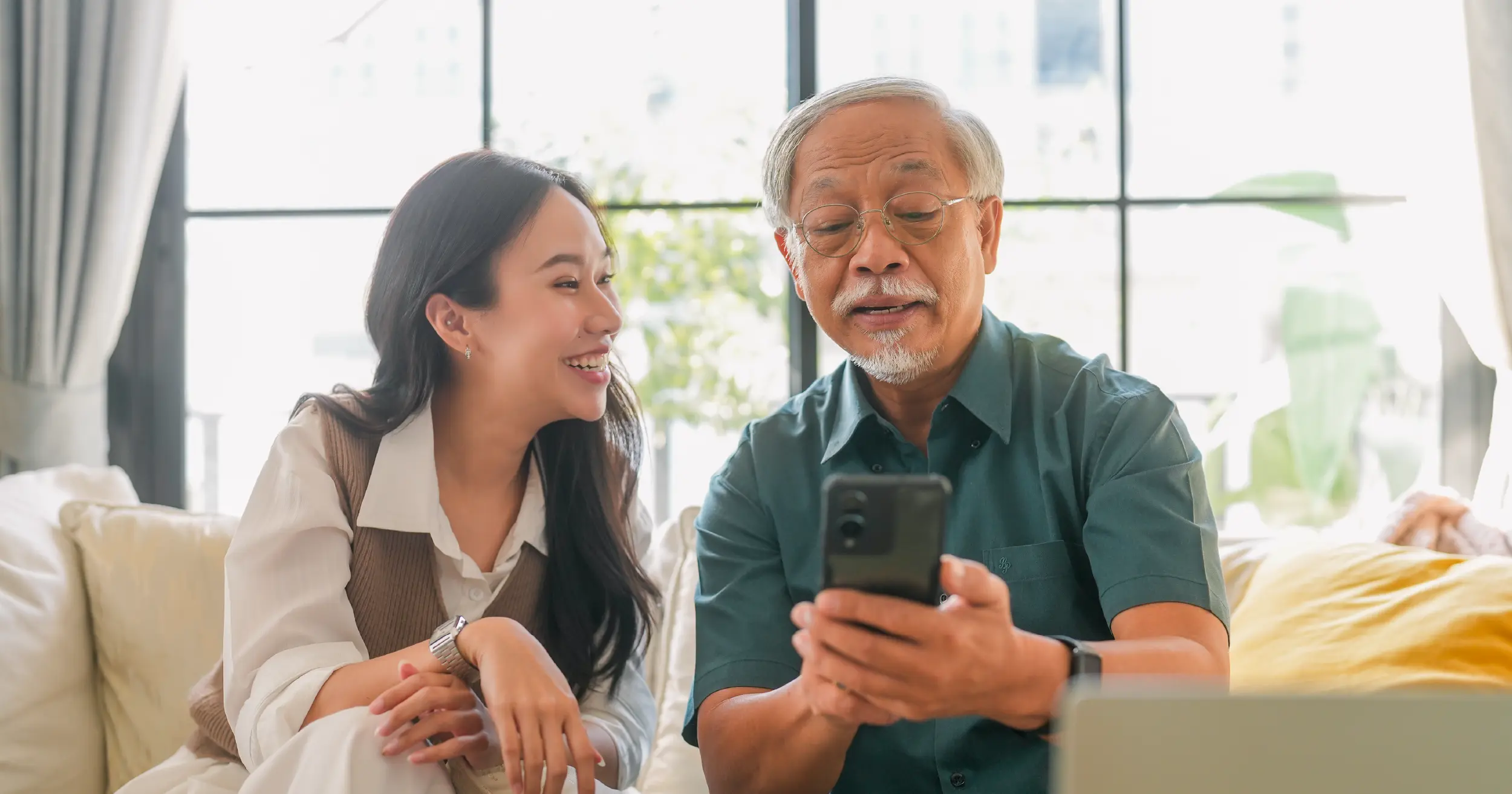 An adult woman smiling at an elderly man as he uses a phone.