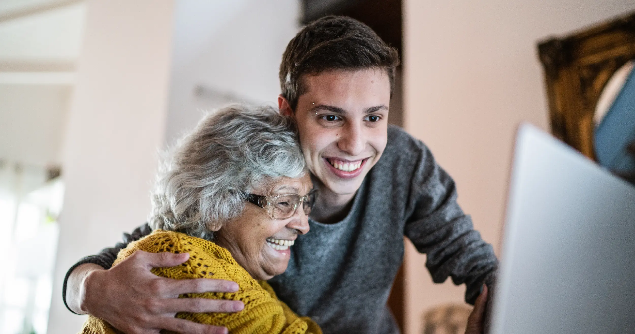A teenage boy hugging an elderly woman as they look at a computer screen, denoting cybersafety.