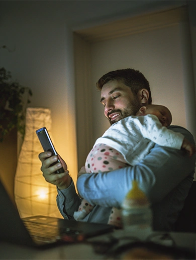 A father smiling as he checks his smartphone, while carrying his child.