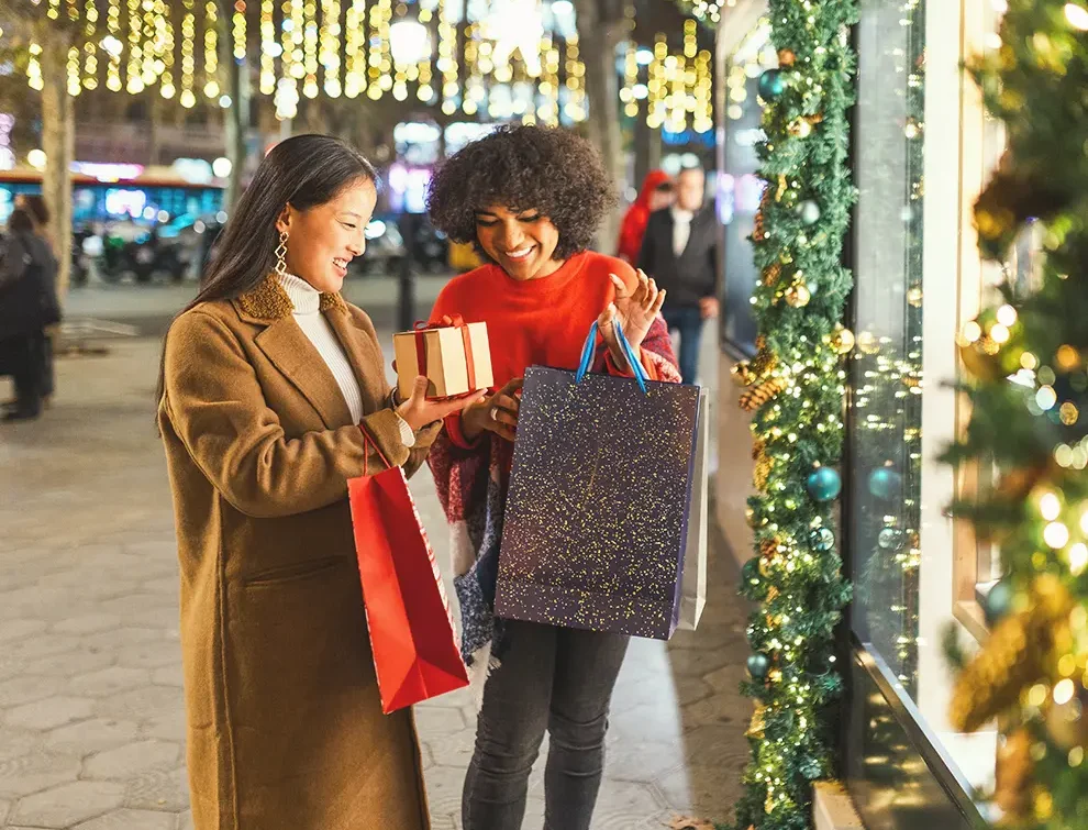 Two women holiday shopping with gift boxes and bags near a decorated store window with Christmas lights.