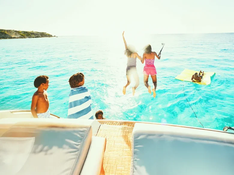 Kids jumping off a boat into clear blue water with others relaxing on a floating mat.